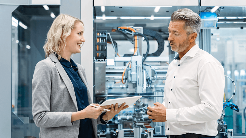 Professionals discussing manufacturing data on tablet in front of automated production system in a modern factory.
