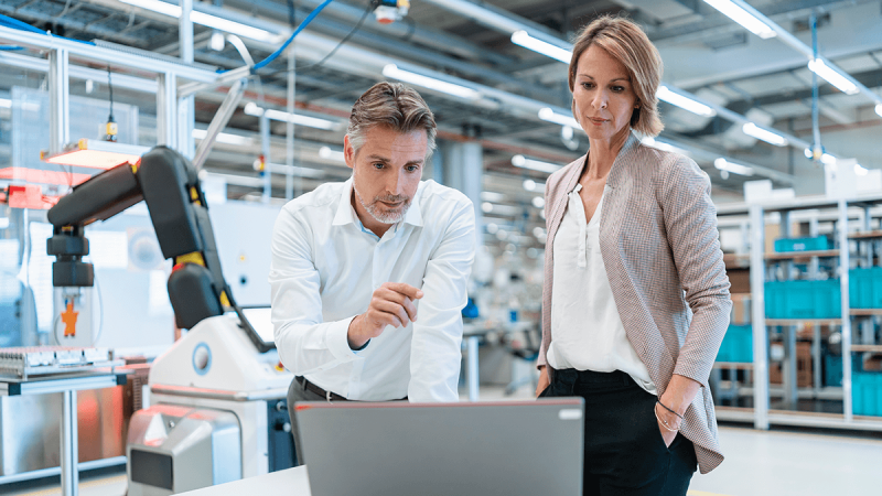 Engineers analyzing production data on laptop in a smart factory environment with industrial robot and automation systems.